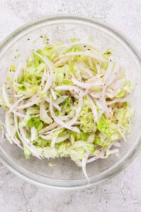 A clear glass bowl containing shredded lettuce and thinly sliced onions mixed with black pepper and seasonings, sitting on a light textured surface.