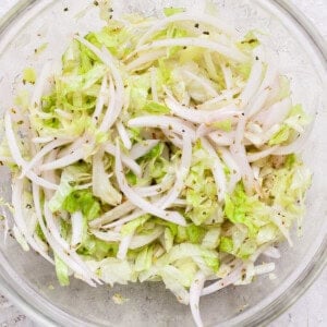 A clear glass bowl containing shredded lettuce and thinly sliced onions mixed with black pepper and seasonings, sitting on a light textured surface.