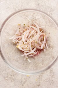 A clear glass bowl containing thinly sliced red onions mixed with dried herbs on a light, textured surface.