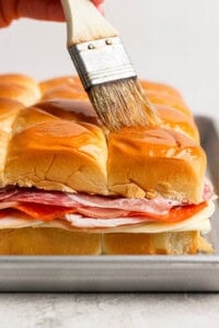 A hand brushes melted butter over the tops of stacked sandwich sliders with layers of deli meats and cheese on a baking tray.
