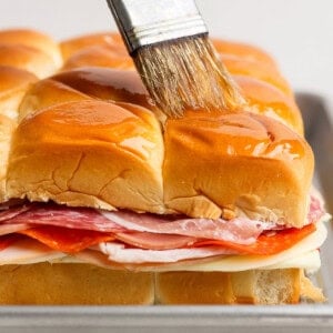 A hand brushes melted butter over the tops of stacked sandwich sliders with layers of deli meats and cheese on a baking tray.