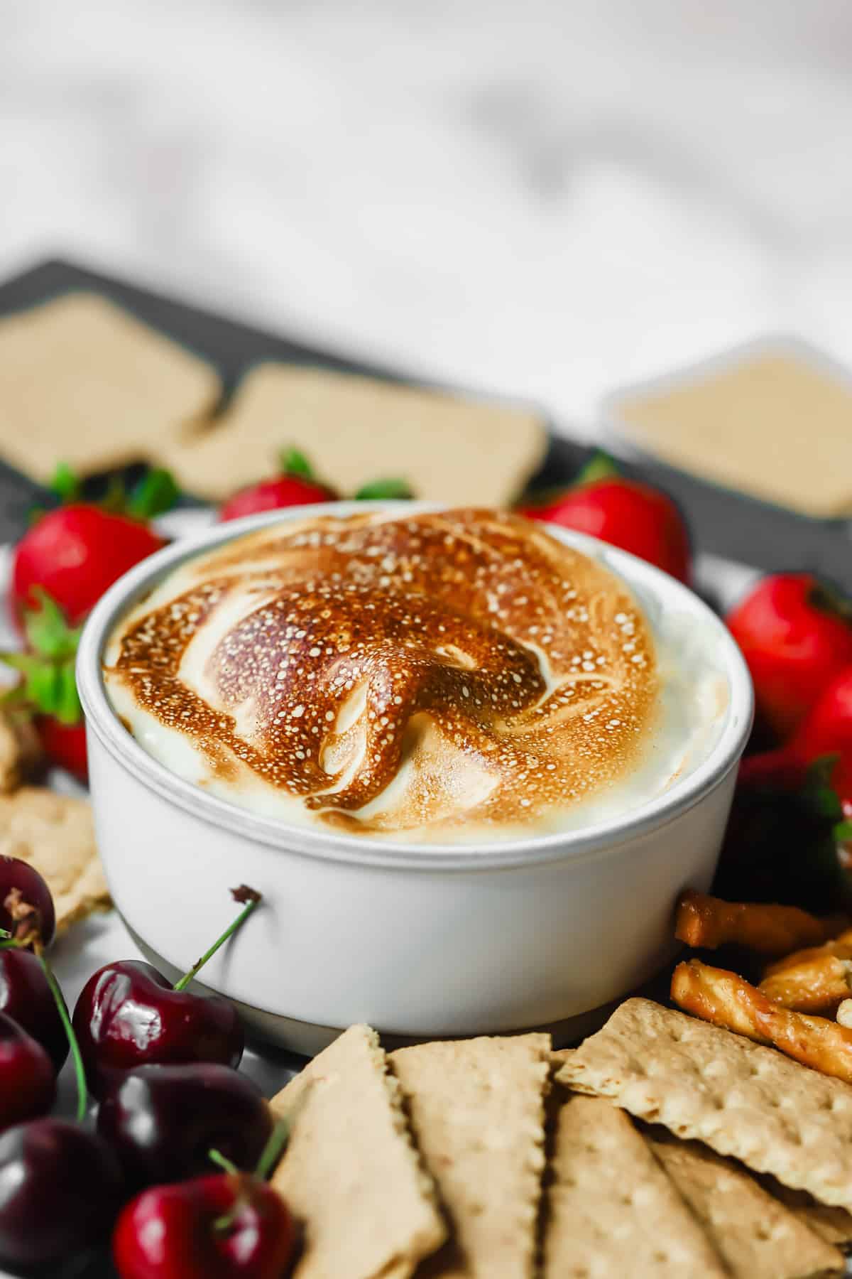 A white bowl filled with toasted marshmallow dip, surrounded by graham crackers, pretzels, strawberries, and cherries.