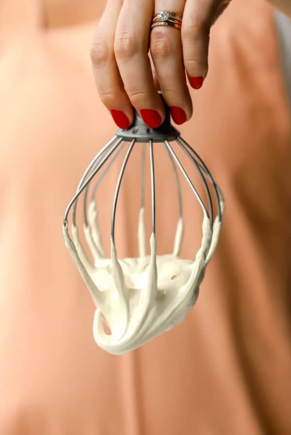 A hand with red nail polish holds a metal whisk coated in thick, creamy batter against a peach-colored background.