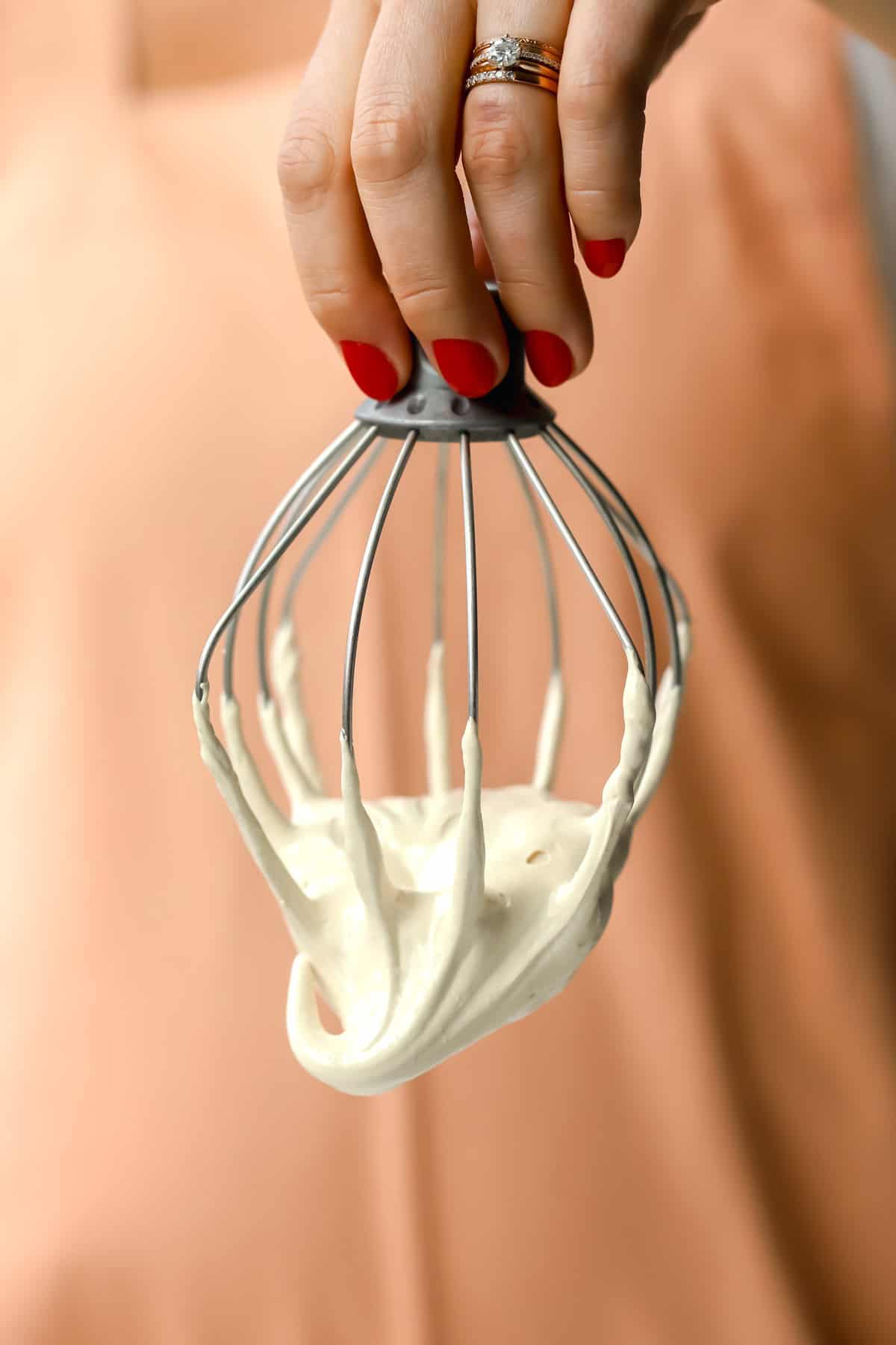A hand with red nail polish holds a metal whisk coated in thick, creamy batter against a peach-colored background.