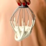 A hand with red nail polish holds a metal whisk covered in thick, maple marshmallow fluff against a peach-colored background.