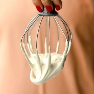 A hand with red nail polish holds a metal whisk covered in thick, maple marshmallow fluff against a peach-colored background.