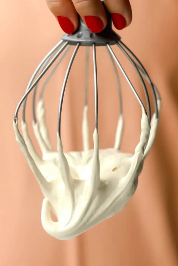 A hand with red nail polish holds a metal whisk covered in thick, maple marshmallow fluff against a peach-colored background.