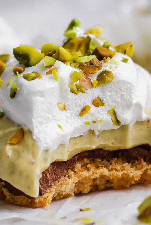A dessert bar with a crumbly base, chocolate layer, creamy filling, whipped topping, and chopped pistachios, with a bite taken out of it.