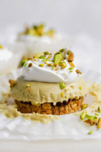 A mini cheesecake with a crumb crust, topped with whipped cream and chopped pistachios, sits on a piece of white parchment paper.