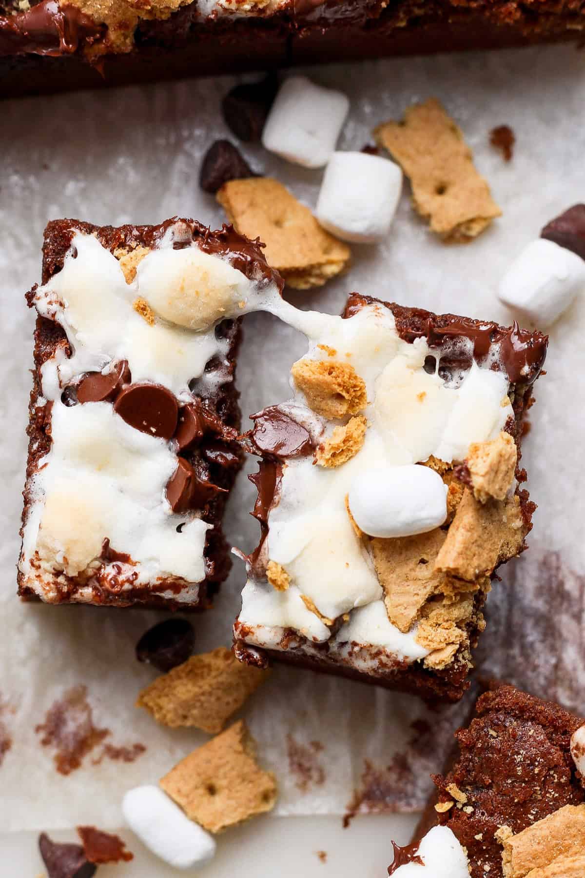 Two s'mores brownies topped with melted marshmallows, chocolate chips, and graham cracker pieces, placed on parchment paper with scattered ingredients around.