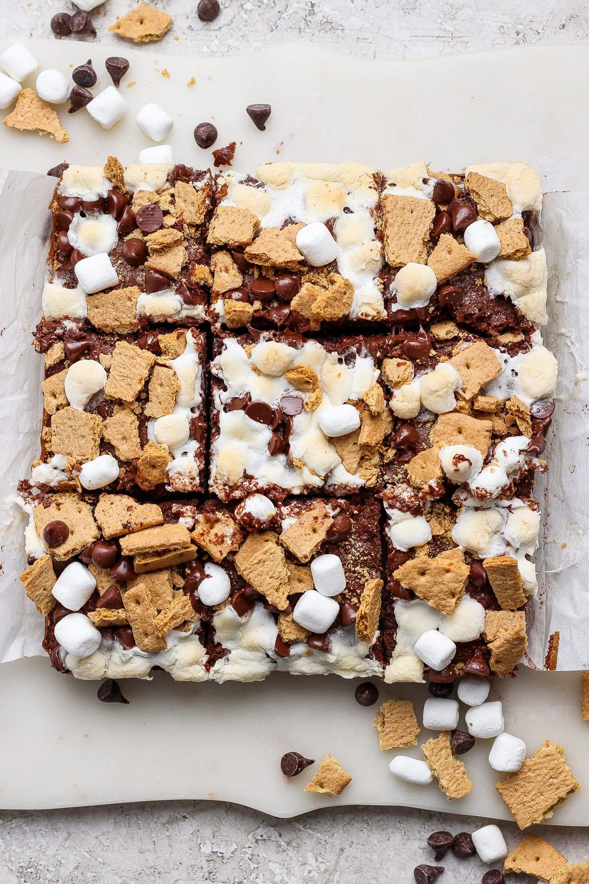 A batch of s'mores bars cut into squares, topped with mini marshmallows, graham cracker pieces, and chocolate chips on parchment paper—these treats are perfect for fans of classic s’mores brownies.