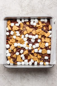 A square baking pan lined with parchment paper, filled with a mixture of mini marshmallows, chocolate chips, and broken graham crackers sits atop a rich s'mores brownies base.