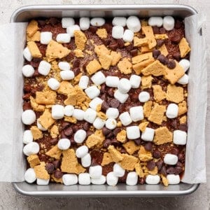 A square baking pan lined with parchment paper, filled with a mixture of mini marshmallows, chocolate chips, and broken graham crackers sits atop a rich s'mores brownies base.