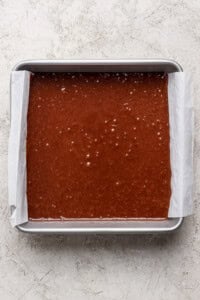 A rectangular metal baking pan lined with parchment paper is filled with s'mores brownies batter on a light textured surface.