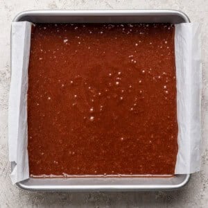 A rectangular metal baking pan lined with parchment paper is filled with s'mores brownies batter on a light textured surface.