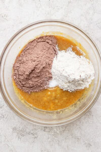 A glass bowl holds a mixture of wet ingredients, with piles of cocoa powder and flour on top, ready to be mixed for delicious s'mores brownies.