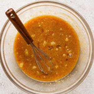 A glass bowl containing a brown, partially mixed s'mores brownies batter with visible lumps and a metal whisk with a wooden handle resting inside.