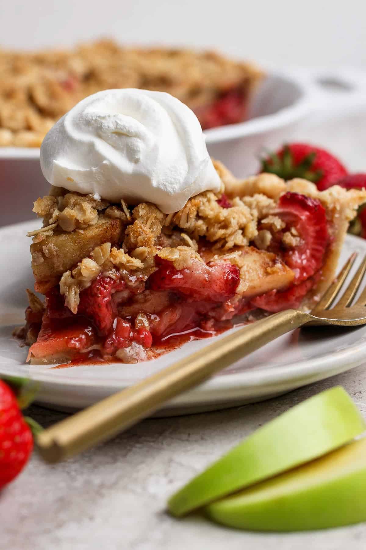 A slice of strawberry apple pie crumble topped with whipped cream sits on a white plate, accompanied by a fork and fresh fruit on the side.