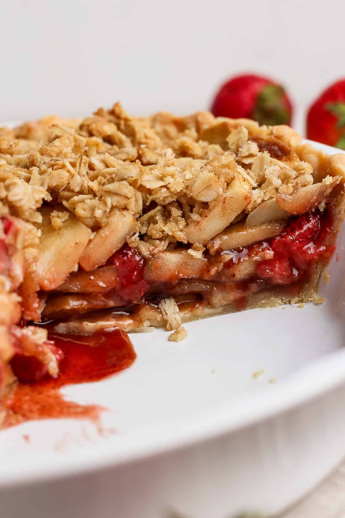 A close-up of a sliced strawberry apple pie crumble with a golden oat topping, filled with layers of apples and strawberries in a white dish.