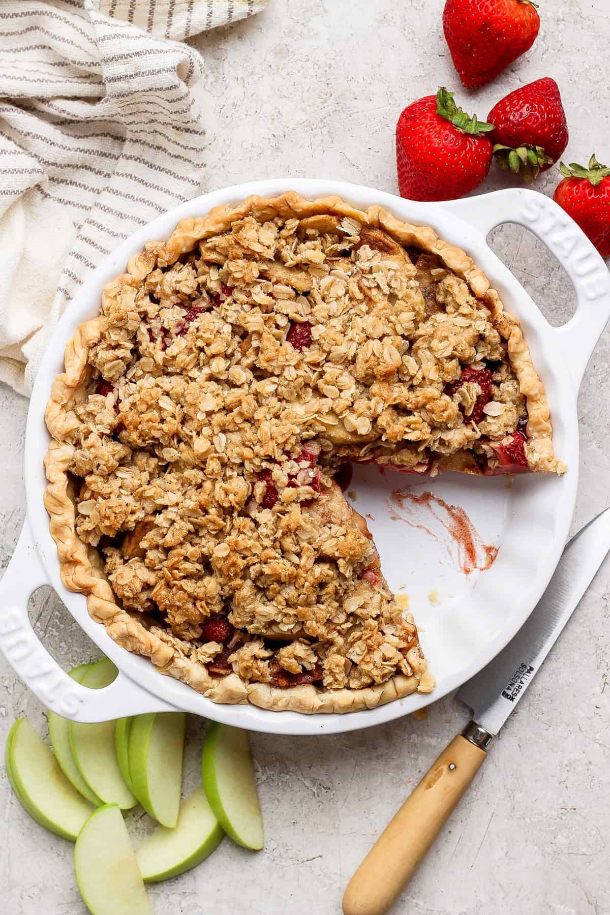 A strawberry apple pie with oat crumble topping sits in a white dish, one slice removed; fresh strawberries, apple slices, and a knife are arranged beside the dish.