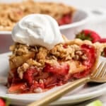 A slice of strawberry apple pie crumble topped with whipped cream sits on a white plate with a gold fork, fresh strawberries in the background.