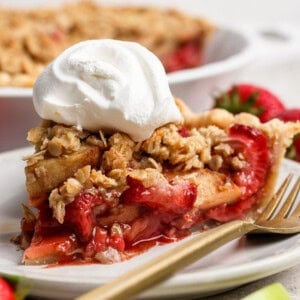 A slice of strawberry apple pie crumble topped with whipped cream sits on a white plate with a gold fork, fresh strawberries in the background.