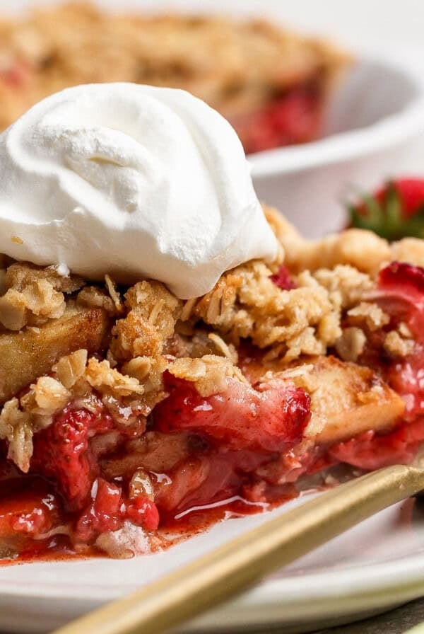A slice of strawberry apple pie crumble topped with whipped cream sits on a white plate with a gold fork, fresh strawberries in the background.
