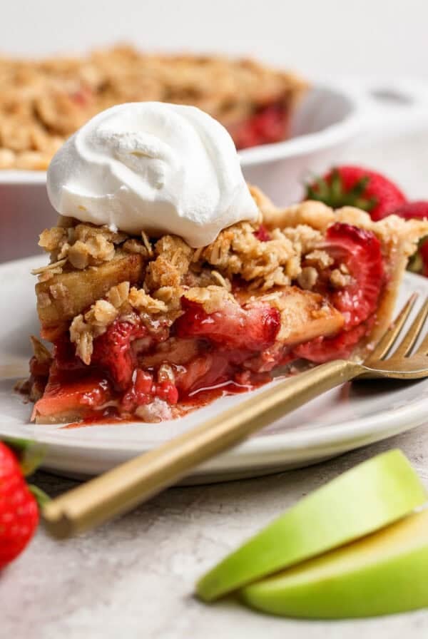 A slice of strawberry apple pie crumble topped with whipped cream sits on a white plate, with a gold fork, fresh strawberries, and apple slices alongside it.