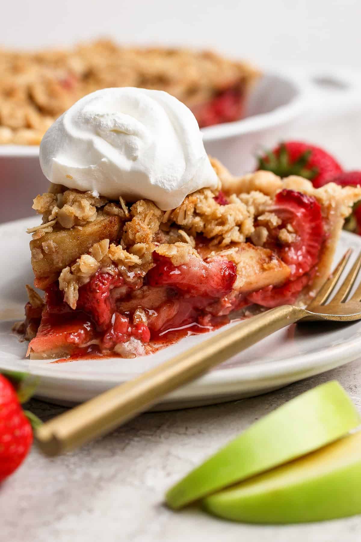 A slice of strawberry apple pie crumble topped with whipped cream sits on a white plate, with a gold fork, fresh strawberries, and apple slices alongside it.