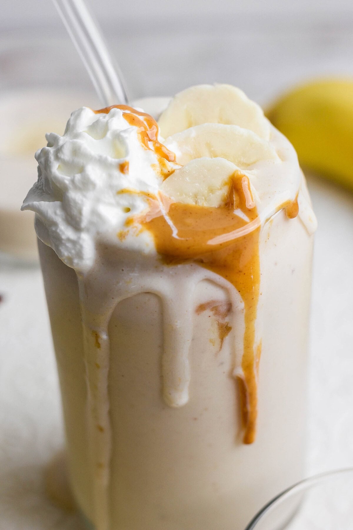 A glass of banana milkshake topped with whipped cream, banana slices, and peanut butter drizzle, with a straw inserted.