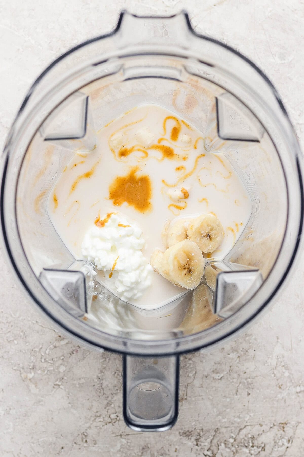 A blender containing milk, sliced banana, cottage cheese, and a drizzle of honey, viewed from above.