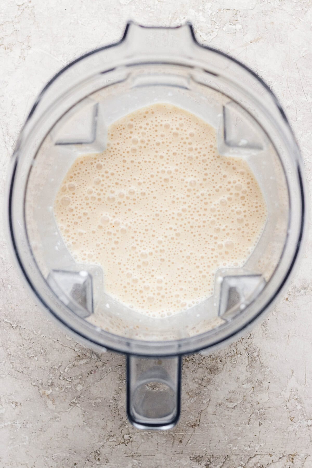 Top-down view of a blender containing a light beige, frothy liquid on a light textured surface.