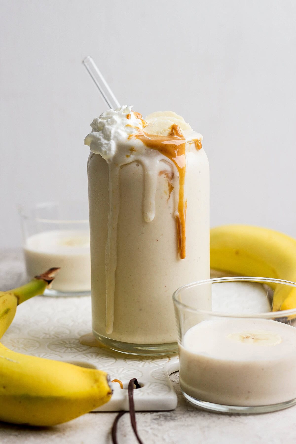 Glass of banana smoothie topped with whipped cream and peanut butter, surrounded by bananas and two small glasses of smoothie.