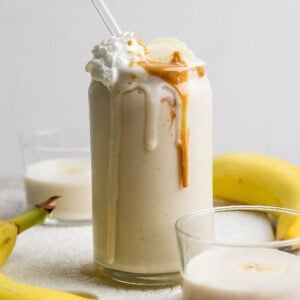 Glass of banana smoothie topped with whipped cream and peanut butter, surrounded by bananas and two small glasses of smoothie.