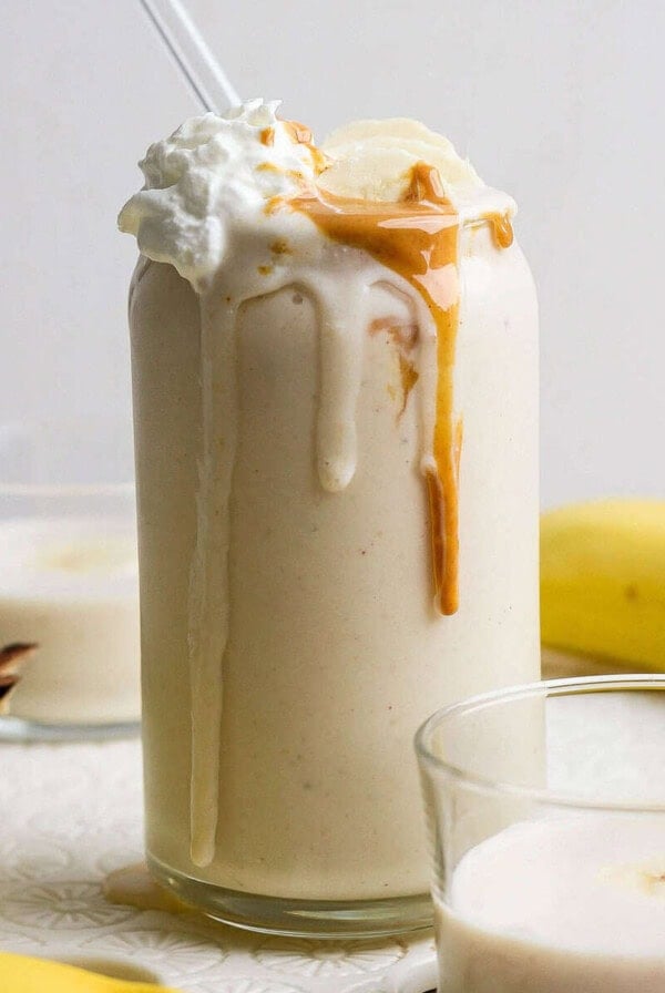 A glass of peanut butter banana protein shake topped with whipped cream, banana slices, and peanut butter, with a straw, surrounded by bananas and two smaller glasses of smoothie.