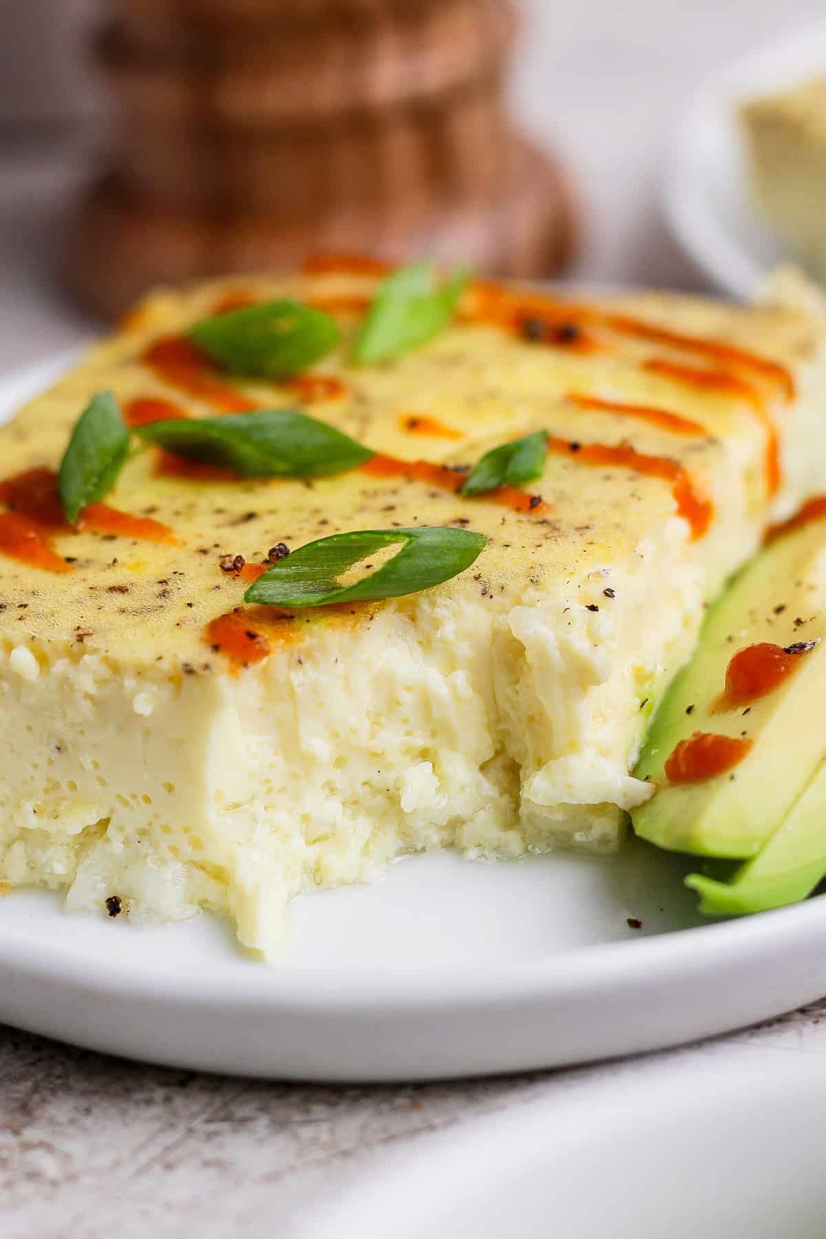 A slice of baked egg casserole topped with sliced green onions and hot sauce, served with sliced avocado on a white plate.