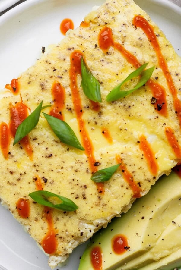 A rectangular egg dish garnished with sliced green onions and hot sauce, served with avocado slices on a white plate.