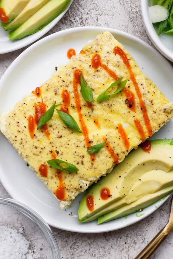 A rectangular egg dish topped with sliced green onions and drizzled with hot sauce, served with avocado slices on a white plate.