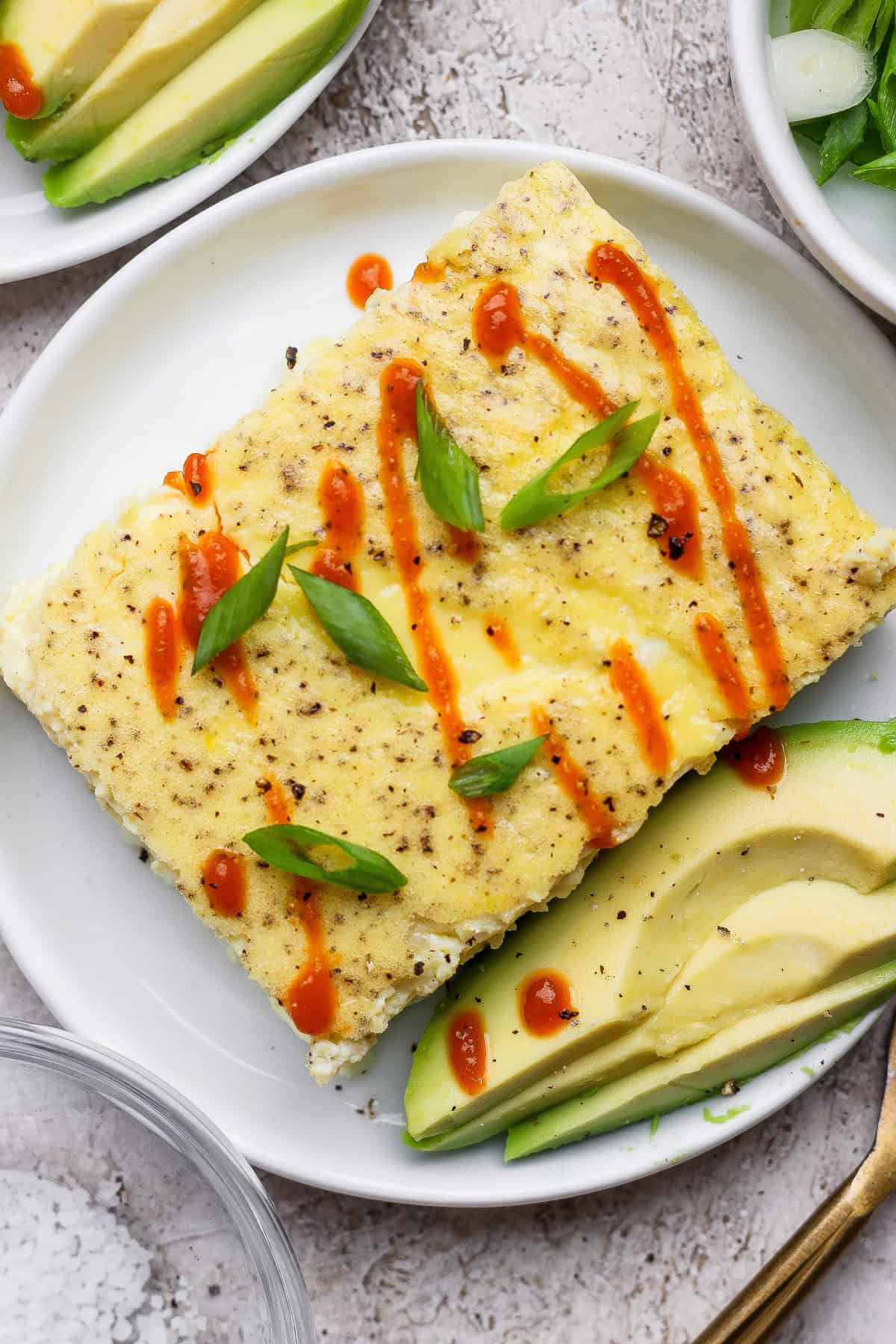 A rectangular egg dish topped with sliced green onions and drizzled with hot sauce, served with avocado slices on a white plate.