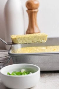 A spatula lifts a rectangular slice of baked egg from a metal pan; a bowl of chopped green onions sits in the foreground.