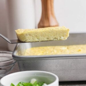 A spatula lifts a rectangular slice of baked egg from a metal pan; a bowl of chopped green onions sits in the foreground.