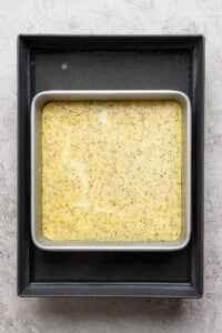 A square metal baking pan filled with a yellow speckled batter sits inside a larger black tray on a textured gray surface.