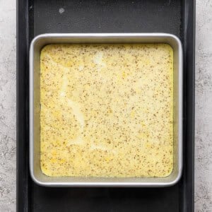 A square metal baking pan filled with a yellow speckled batter sits inside a larger black tray on a textured gray surface.