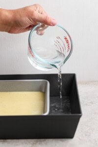 A hand pours water from a glass measuring cup into a larger baking pan, which holds a smaller pan filled with batter.