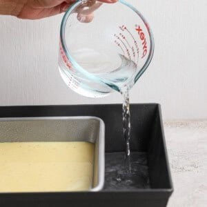 A hand pours water from a glass measuring cup into a larger baking pan, which holds a smaller pan filled with batter.
