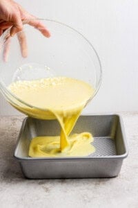 A hand pours yellow cake batter from a glass bowl into a square metal baking pan on a countertop.