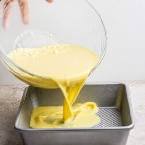 A hand pours yellow cake batter from a glass bowl into a square metal baking pan on a countertop.