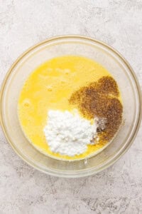 A glass bowl containing whisked eggs, cottage cheese, ground black pepper, and salt on a light textured surface.