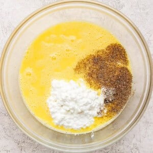 A glass bowl containing whisked eggs, cottage cheese, ground black pepper, and salt on a light textured surface.