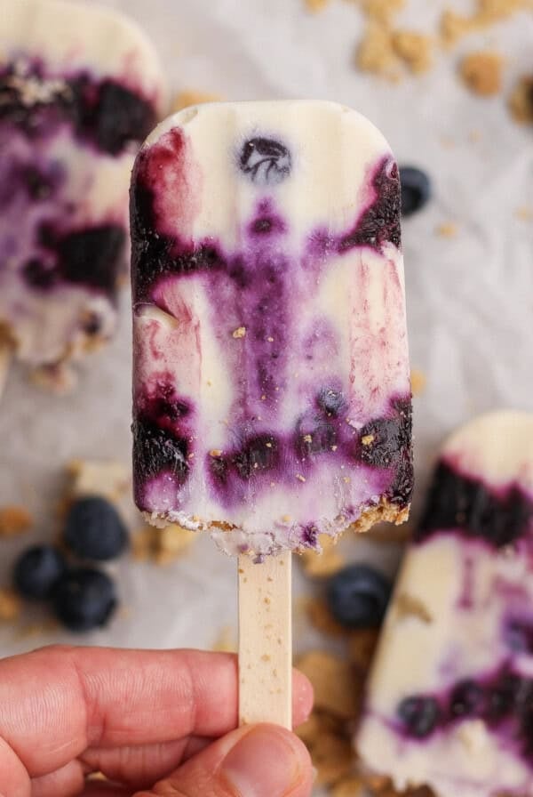 A hand holds a partially eaten blueberry swirl yogurt popsicle on a stick, with blueberries and crumbs scattered in the background.